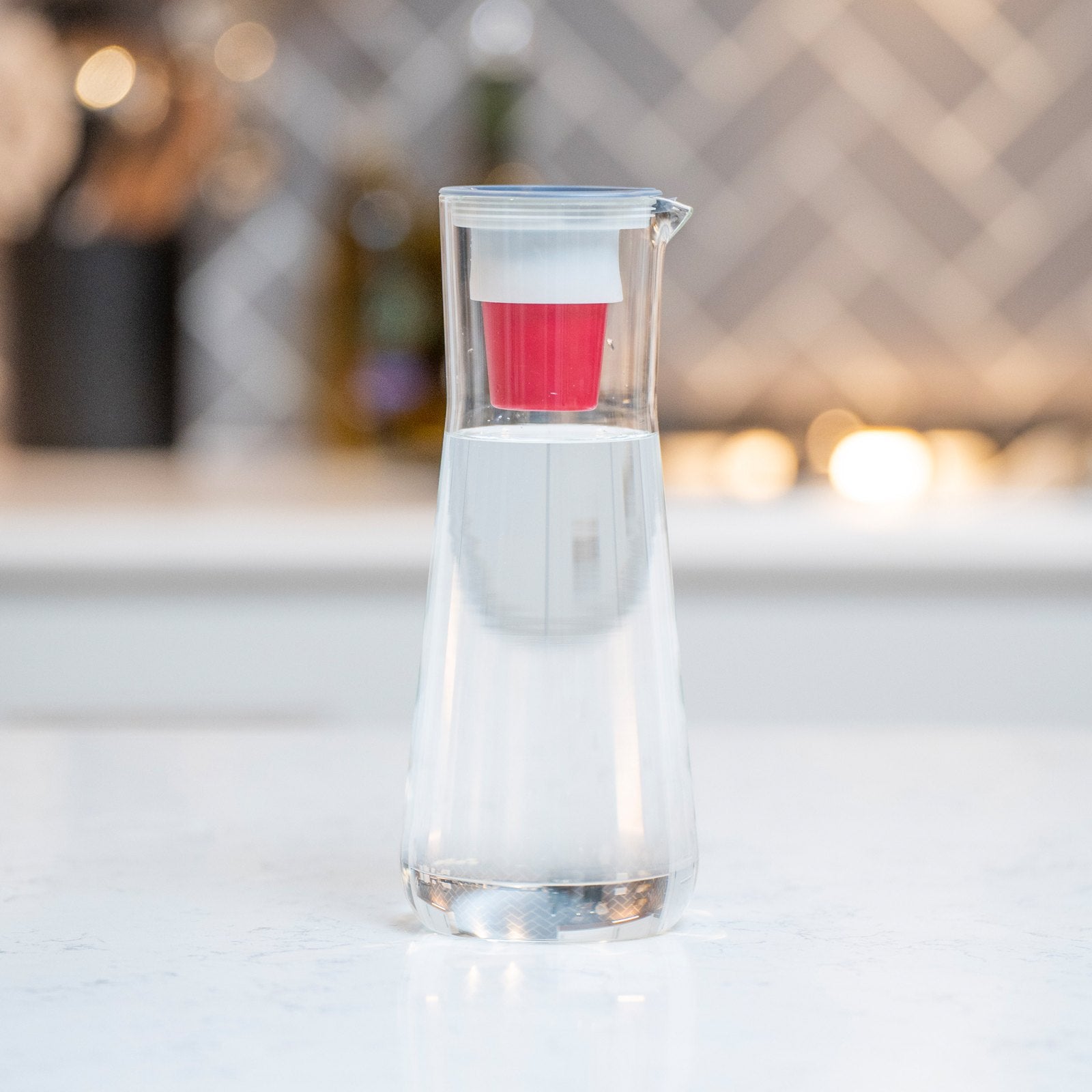 Clear narrow Hydros glass pitcher holding water with its red filter visible in the lid